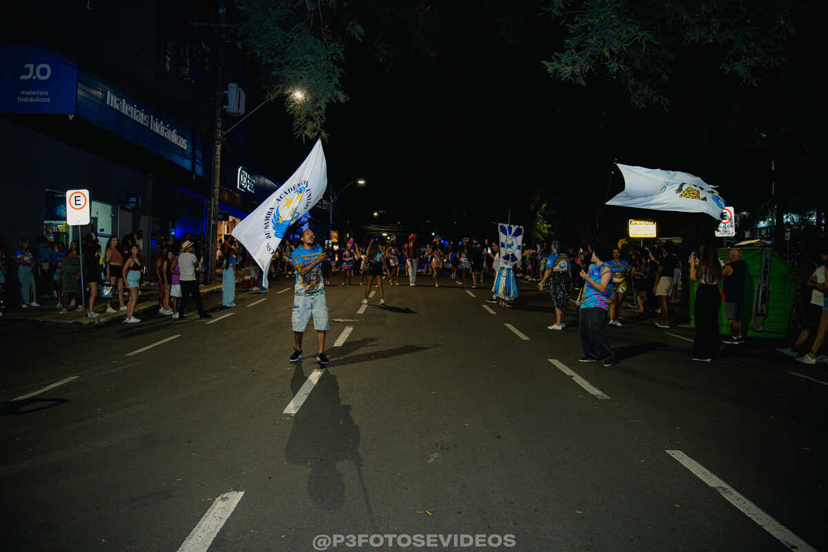 Integrantes da Escola de Samba SCB União desfilando à noite em rua de Santa Cruz do Sul com bandeiras brancas.